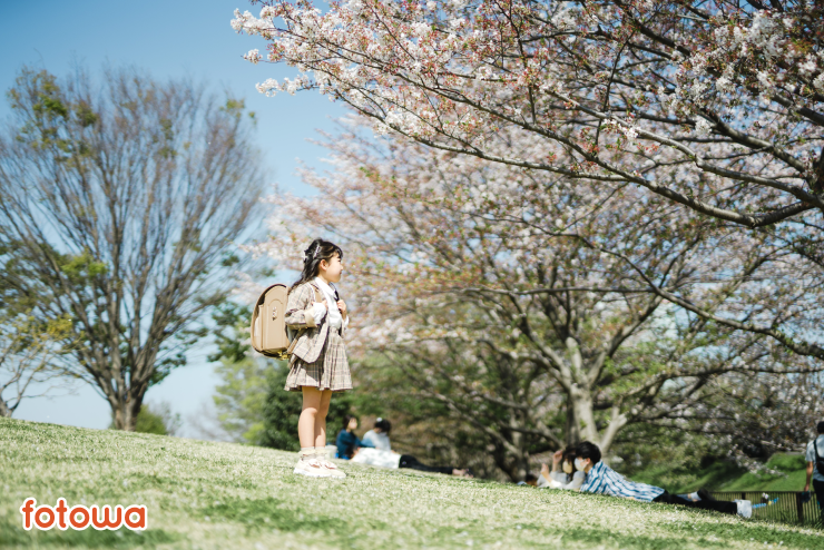 桜の木の横に立つベージュのランドセルを背負った女の子