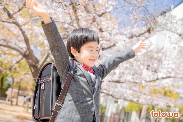 満開の桜の前で両手を挙げて微笑むランドセルを背負った男の子