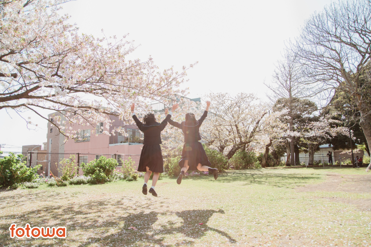桜の木の横で両手をあげて高くジャンルする制服を着た2人の女の子